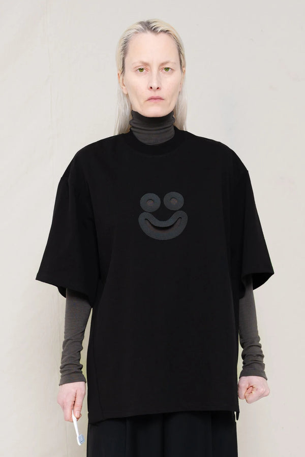 PERSON-WITH-GREEN-EYES-WEARING-BLACK-OVERSIZE-HAPPY-SMILEY-SHIRT-OVER-GREY-TURTLENECK-LONGSLEEVE-AND-WEARS-BLACK-WIDE-TROUSERS-AND-MUD-CUSTOMIZED-NIKE-SNEAKER-IN-GREY-HOLDING-TOOTHBRUSH-FRONT-VIEW