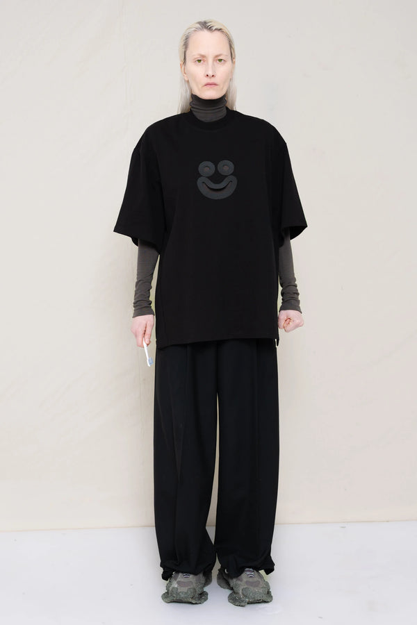 woman-holding-a-tooth-brush-wearing-a-black-oversized-tee-with-a-smiley-face-cut-out-over-a-long-sleeve-grey-turtle-neck-and-black-WOOL-BLEND-TWILL-TAILORED-PANTS-WITH-ZIGZAG-HEM-DETAIL-worn-with-custom-shoes-full-body-front-view
