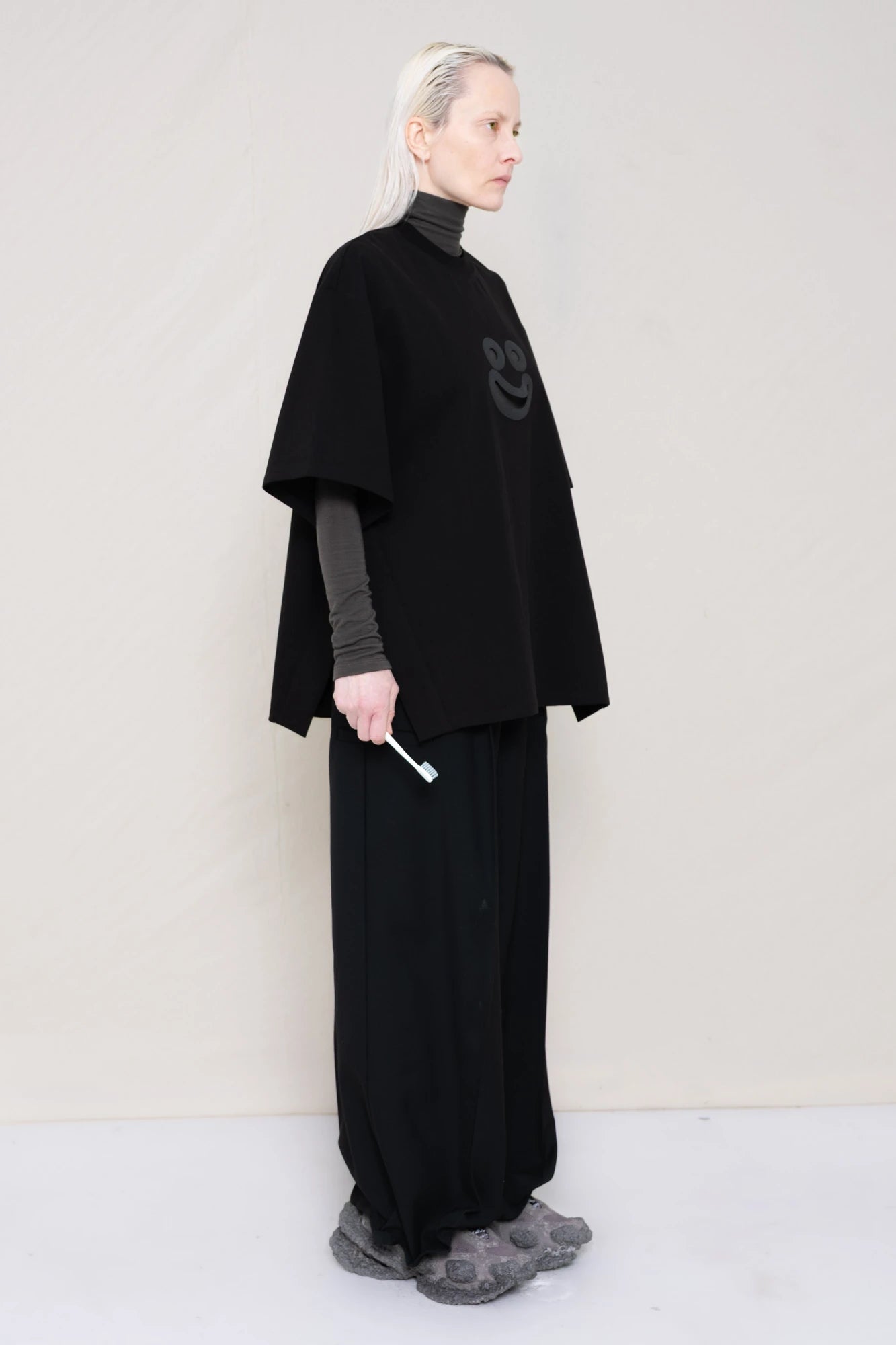 PERSON-WITH-GREEN-EYES-WEARING-BLACK-OVERSIZE-HAPPY-SMILEY-SHIRT-OVER-GREY-TURTLENECK-LONGSLEEVE-AND-WEARS-BLACK-WIDE-TROUSERS-AND-MUD-CUSTOMIZED-NIKE-SNEAKER-IN-GREY-HOLDING-TOOTHBRUSH-QUATER-FRONT-VIEW