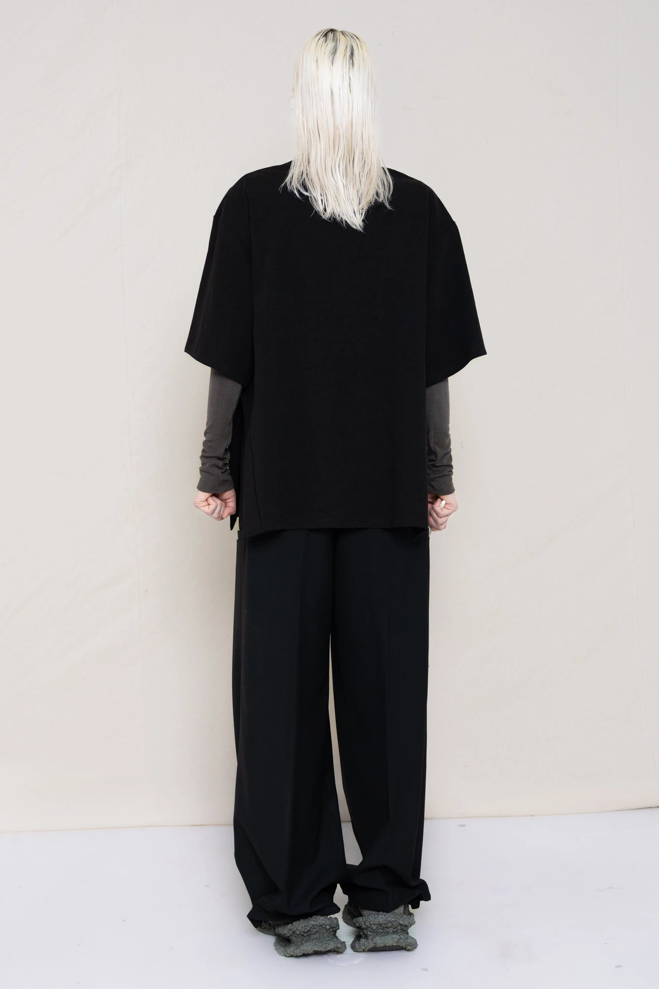 PERSON-WITH-GREEN-EYES-WEARING-BLACK-OVERSIZE-HAPPY-SMILEY-SHIRT-OVER-GREY-TURTLENECK-LONGSLEEVE-AND-WEARS-BLACK-WIDE-TROUSERS-AND-MUD-CUSTOMIZED-NIKE-SNEAKER-IN-GREY-HOLDING-TOOTHBRUSH-BACK-VIEW