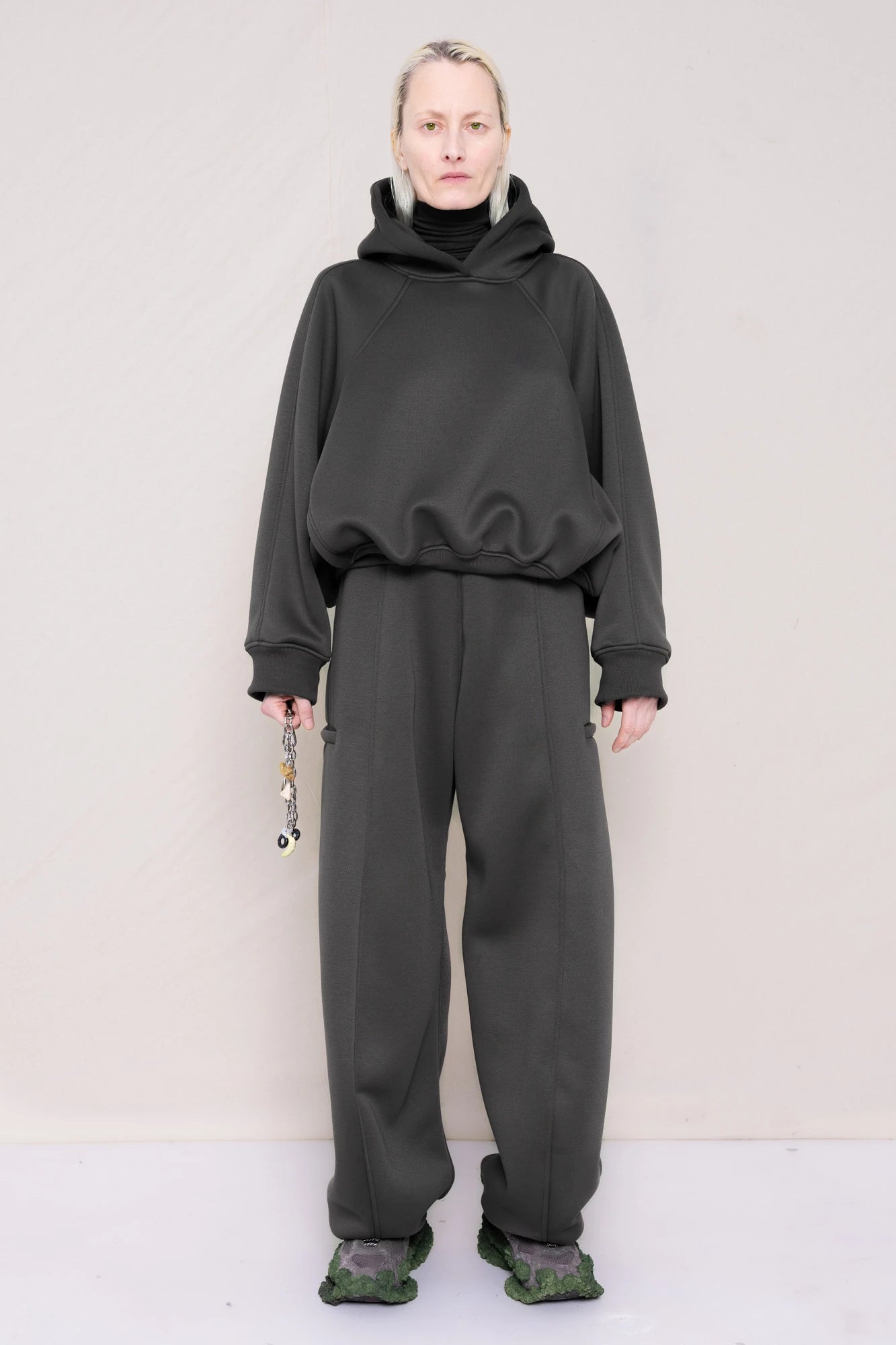PERSON-WEARING-STONE-GREY-CROPPED-HOODIE-WITH-MATCHING-STONE-GREY-ROUND-LEG-LOUNGEPANTS-HOLDING-MB-CHARM-NECKLACE-WITH-CUSTOMIZED-SNEAKERS-FRONT-VIEW