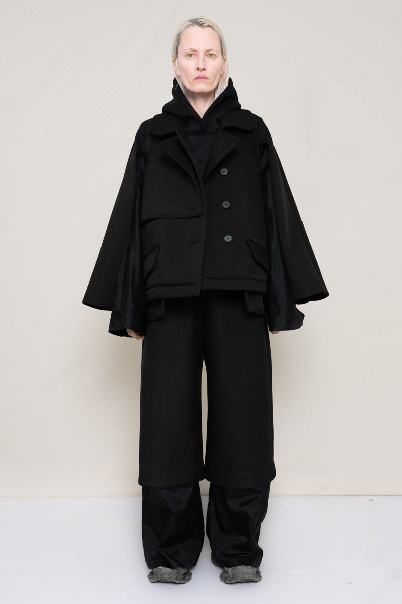A-model-wearing-A-black-DOUBLE-LAPEL-JACKET-WITH-black-SILKY-SECOND-SLEEVEs-over-a-spongey-black-hoodie-and-black-trousers-with-a-black-silky-underlayer-draped-from-under-the-cuffs-AND-CUSTOMIZED-SNEAKERS-full-body-front-view