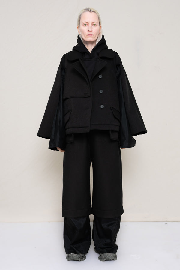 A-model-wearing-A-black-DOUBLE-LAPEL-JACKET-WITH-black-SILKY-SECOND-SLEEVEs-over-a-spongey-black-hoodie-and-black-trousers-with-a-black-silky-underlayer-draped-from-under-the-cuffs-AND-CUSTOMIZED-SNEAKERS-full-body-front-view