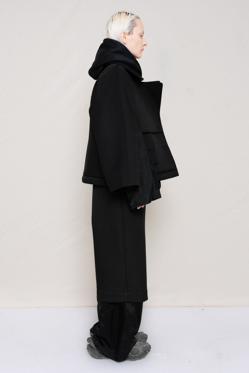 A-model-wearing-A-black-DOUBLE-LAPEL-JACKET-WITH-black-SILKY-SECOND-SLEEVEs-over-a-spongey-black-hoodie-and-black-trousers-with-a-black-silky-underlayer-draped-from-under-the-cuffs-AND-CUSTOMIZED-SNEAKERS-full-body-side-view