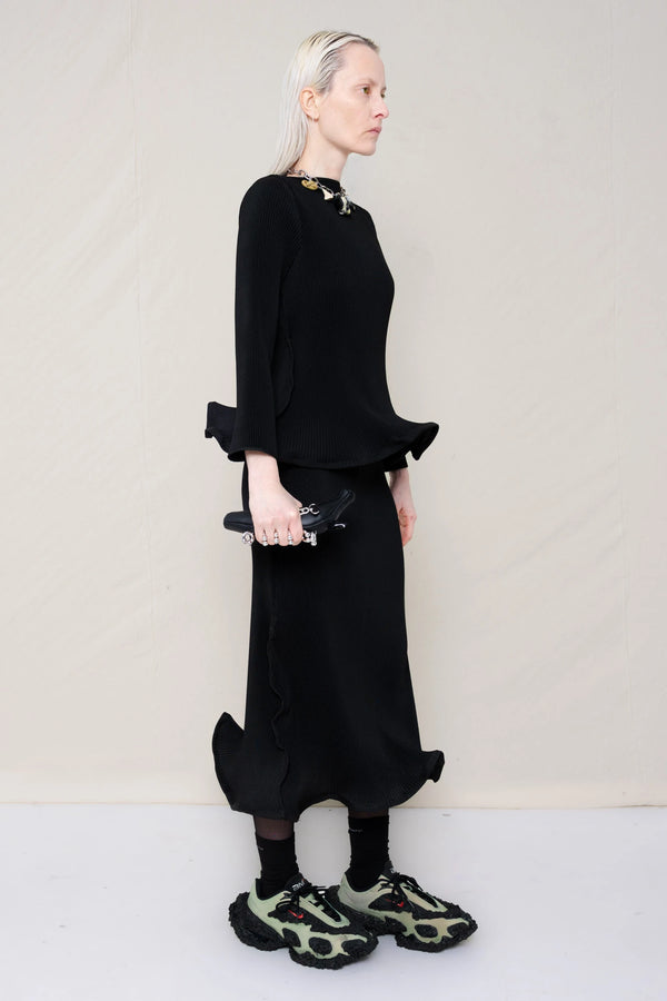 PERSON-WEARING-BLACK-RIPPLE-PLEAT-ZIG-ZAG-HEM-TOP-AND-MATCHING-RIPPLE-PLEAT-ZIG-ZAG-HEM-SKIRT-ACCESSORIZED-WITH-CUSTOMIZED-SNEAKERS-AND-MB-CHARM-NECKLACE-HOLDING-BLACK-PIERCED-BANANA-TOTE-THREE-QUARTER-VIEW