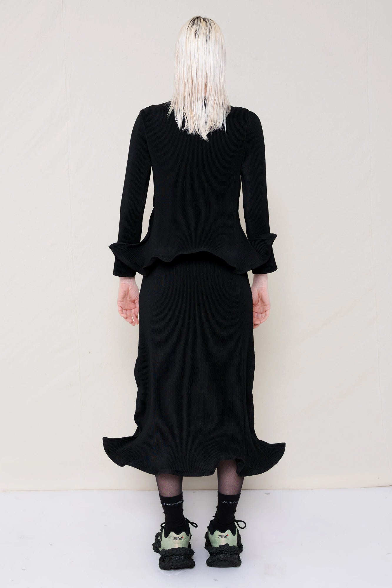 PERSON-WEARING-BLACK-RIPPLE-PLEAT-ZIG-ZAG-HEM-TOP-AND-MATCHING-RIPPLE-PLEAT-ZIG-ZAG-HEM-SKIRT-ACCESSORIZED-WITH-CUSTOMIZED-SNEAKERS-BACK-VIEW