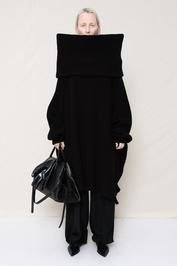 PERSON-WITH-GREEN-EYES-WEARING-BLACK-OVERSIZE-LONG-ROLL-NECK-OVER-SHOULDERS-SWEATER-WITH-BLACK-LONG-TAILORED-TROUSERS-AND-BLACK-COWHIDE-BOOTS-HOLDING-BLACK-A-LINE-SHOPPER-WITH-GLOVES-DETAILS-FRONT-VIEW