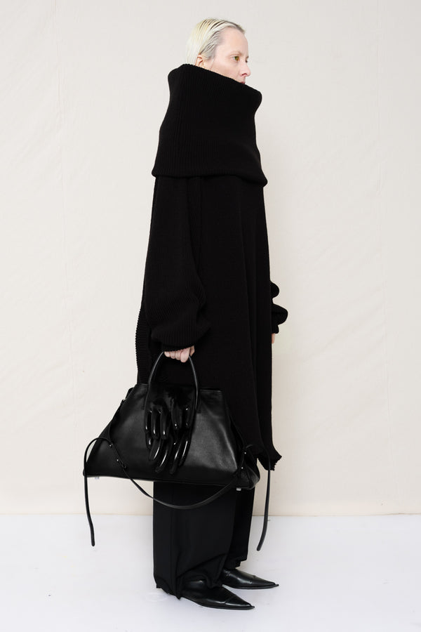 PERSON-WITH-GREEN-EYES-WEARING-BLACK-OVERSIZE-LONG-ROLL-NECK-SWEATER-WITH-BLACK-LONG-TAILORED-TROUSERS-AND-BLACK-COWHIDE-BOOTS-HOLDING-BLACK-A-LINE-SHOPPER-WITH-GLOVE-DETAILS-QUATER-FRONT-VIEW