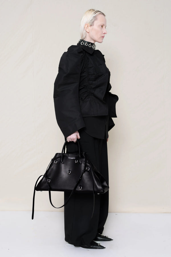 PERSON-WEARING-BLACK-GATHERING-SHIRT-OVER-BLACK-SHIRT-THROW-OVER-WITH-BLACK-LONG-TAILORED-TROUSERS-AND-BLACK-COWHIDE-FRONT-PIERCED-BOOTS-HOLDING-BLACK-A-LINE-SHOPPER-WITH-PIERCING-DETAILS-AND-WEAR-ACCESOIRES-WITH-ALLOVER-PIERCED-NECKLACE-FRONT-QUATER-VIEW