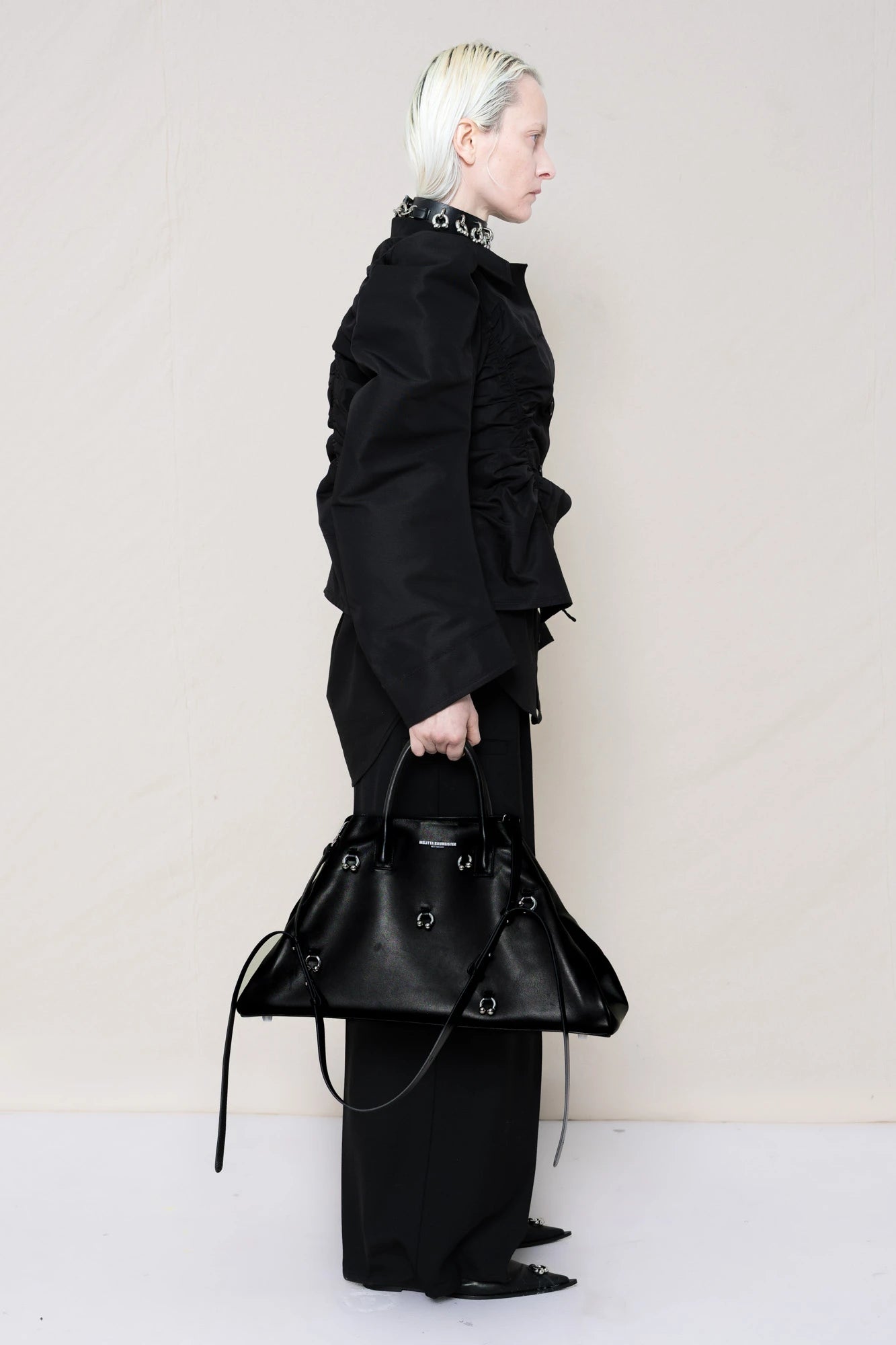 PERSON-WEARING-BLACK-GATHERING-SHIRT-OVER-BLACK-SHIRT-THROW-OVER-WITH-BLACK-LONG-TAILORED-TROUSERS-AND-BLACK-COWHIDE-FRONT-PIERCED-BOOTS-HOLDING-BLACK-A-LINE-SHOPPER-WITH-PIERCING-DETAILS-AND-WEAR-ACCESOIRES-WITH-ALLOVER-PIERCED-NECKLACE-SIDE-VIEW