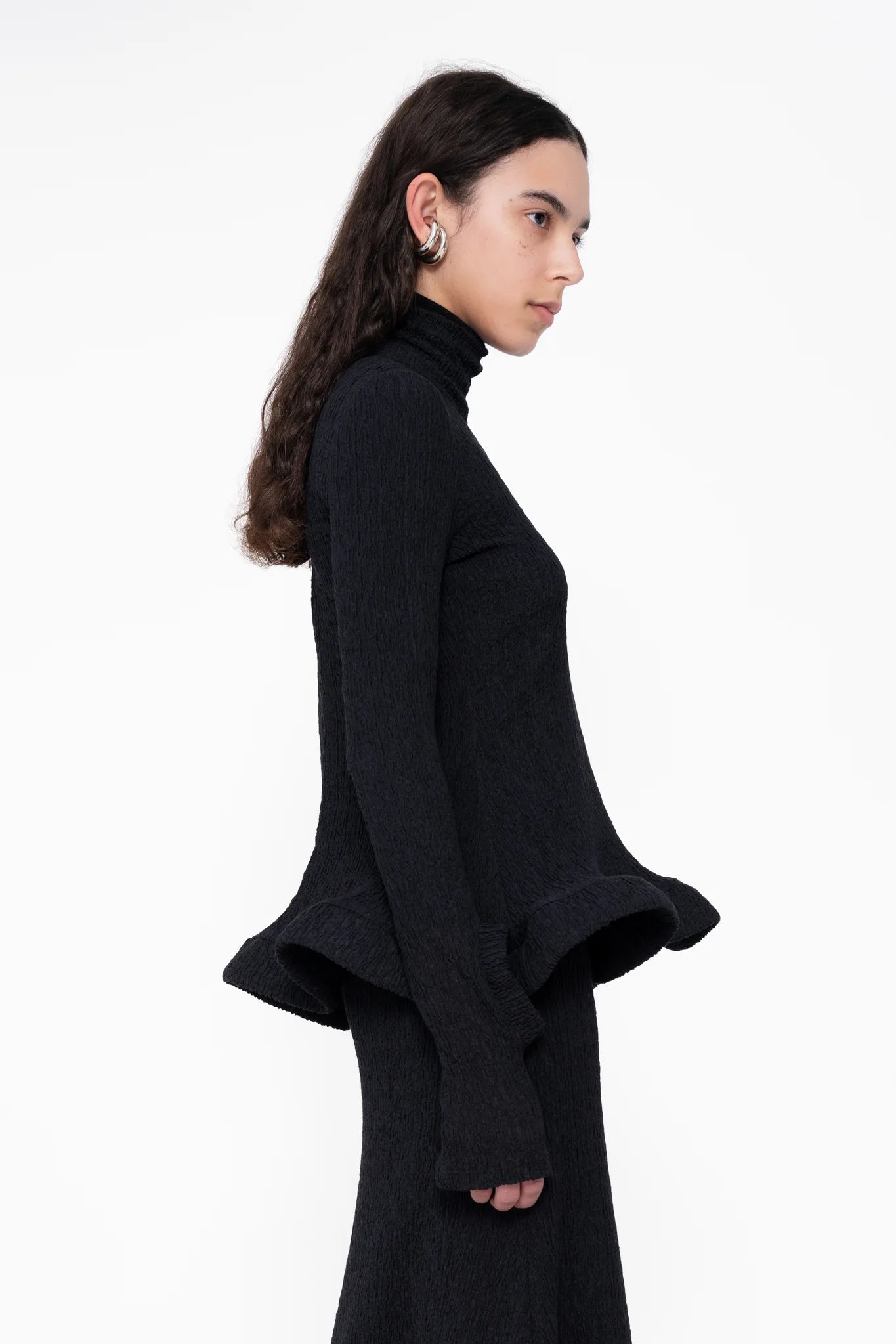 female-wearing-tight-black-longsleeve-turtleneck-top-with-wavy-hem-and-hole-in-sleeve-detail-on-white-background-close-up-torso-side