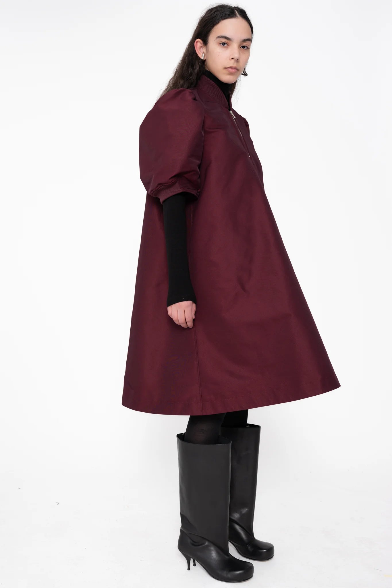 female-wearing-bordeaux-midi-bomber-dress-with-zip-and-cropped-puffed-sleeves-with-black-underlayer-and-boots-on-white-background-full-body-side