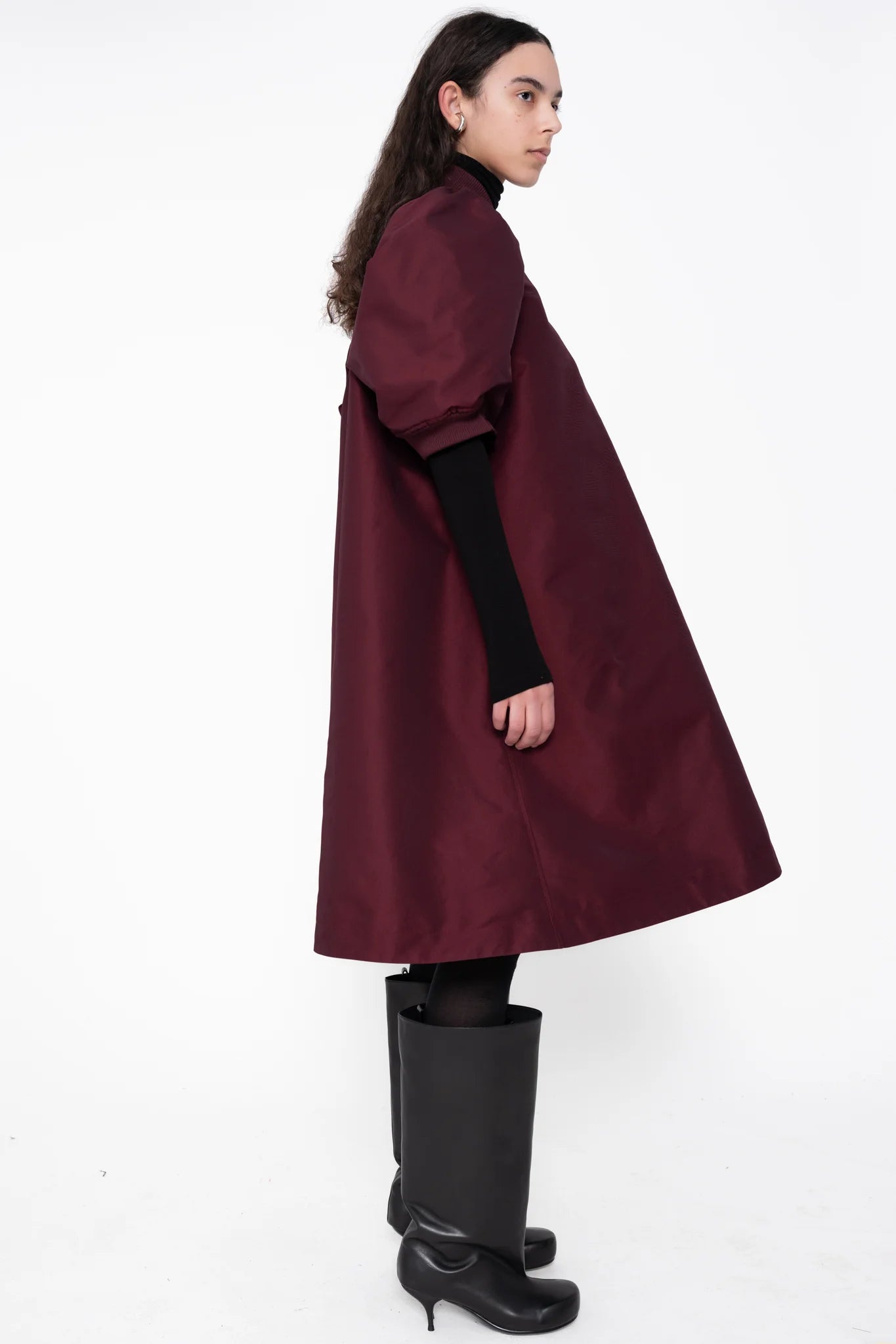 female-wearing-bordeaux-midi-bomber-dress-with-zip-and-cropped-puffed-sleeves-with-black-underlayer-and-boots-on-white-background-full-body-side