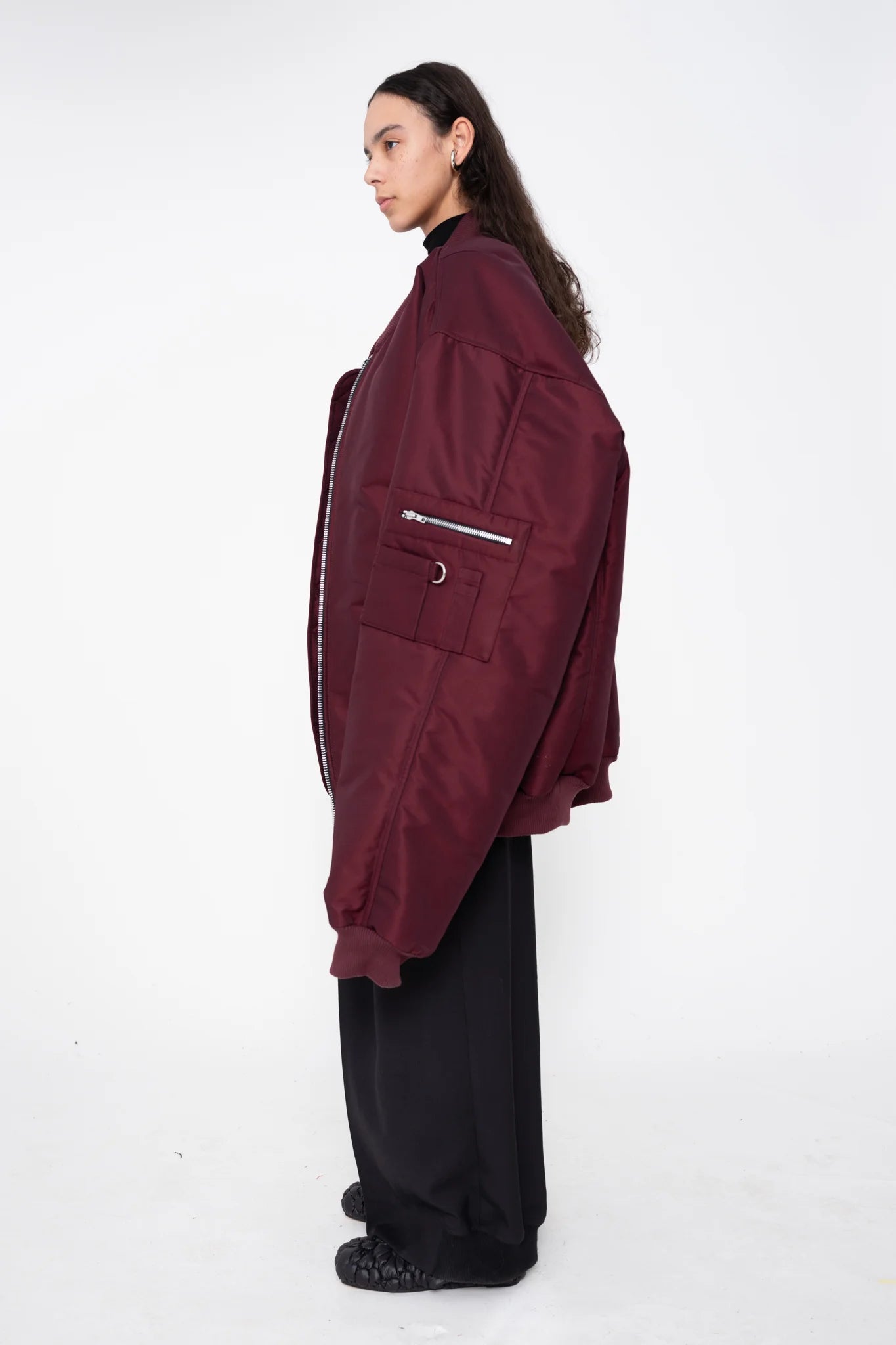 female-wearing-oversized-bordeaux-bomber-jacket-with-slits-at-armholes-and-black-underlayer-top-with-black-suit-pants-on-white-background-full-body-side