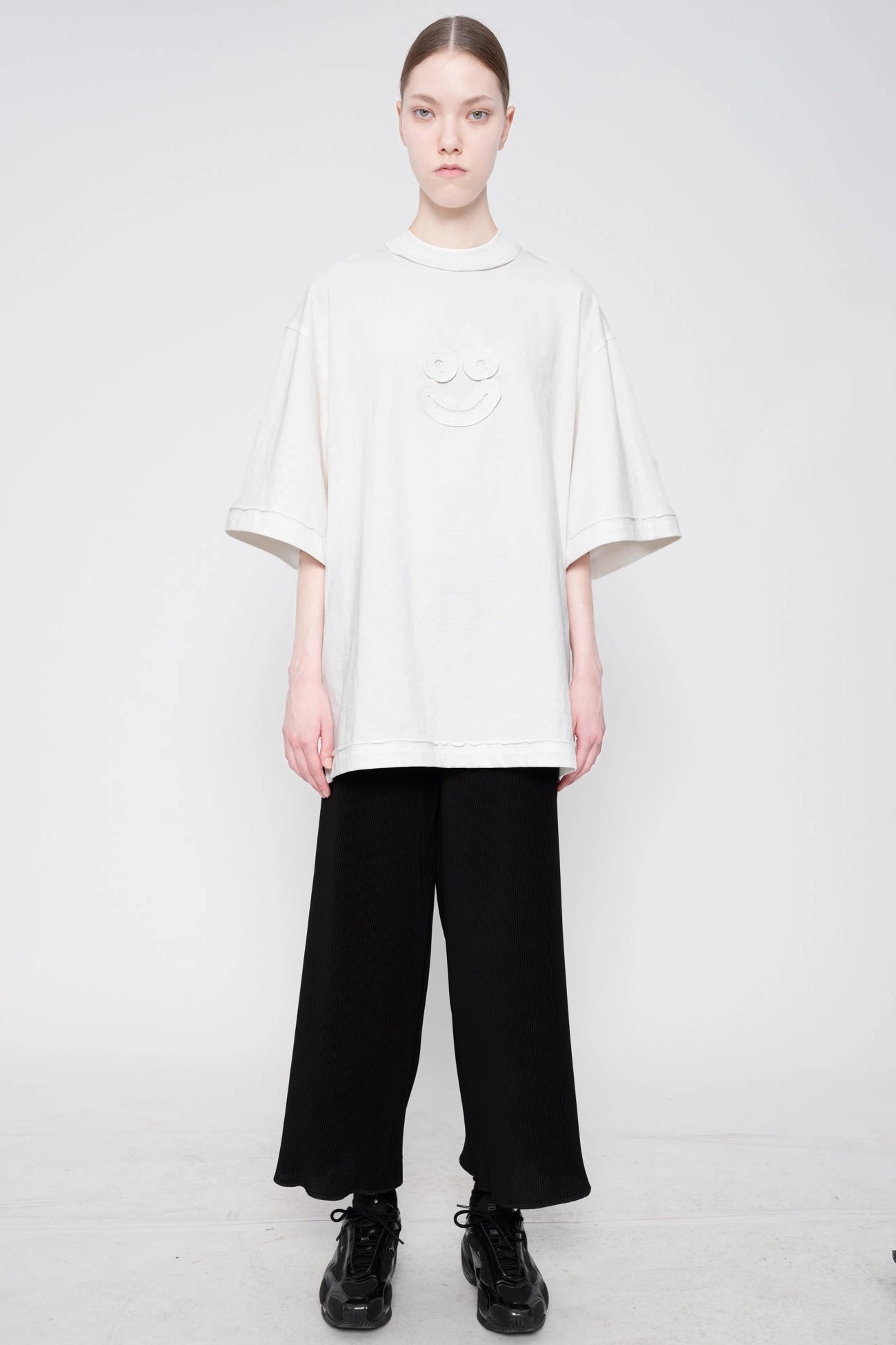 A model stands against a light gray studio background wearing an oversized off-white short-sleeve t-shirt with a subtle smiley face detail on the chest, paired with black wide-leg trousers and black lace-up shoes.