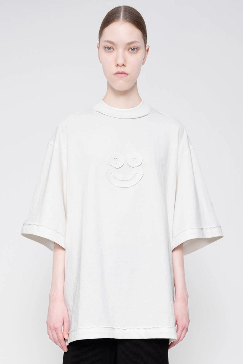 A model wearing an oversized off-white short-sleeve t-shirt with a tonal embroidered smiley face on the chest, set against a plain light gray background.