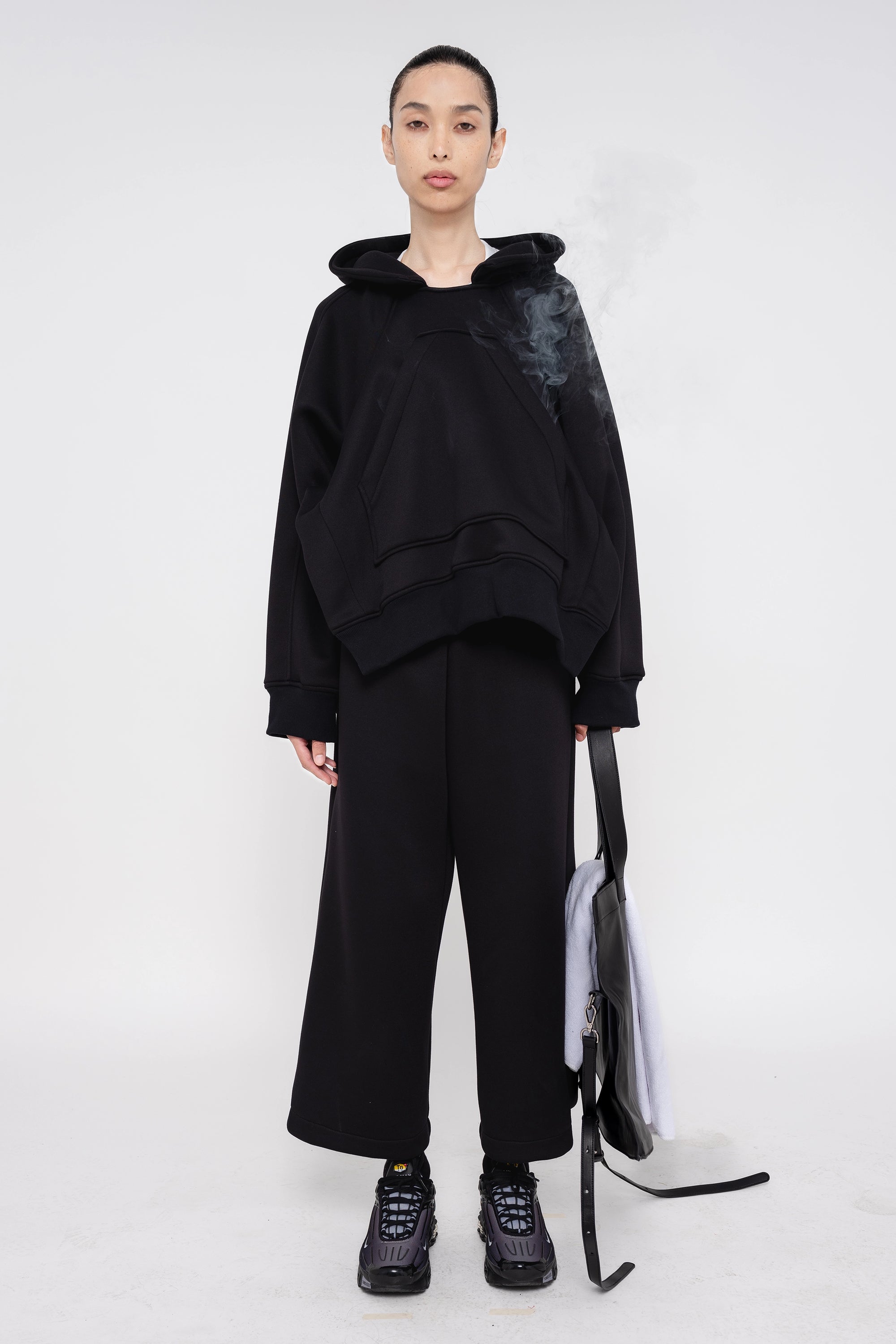 brunette-model-white-background-wearing-black-cropped-hoodie-with-black-lounge-pants-and-sneakers-while-holding-black-slouchy-bag-and-white-banana-tote-full-body-front