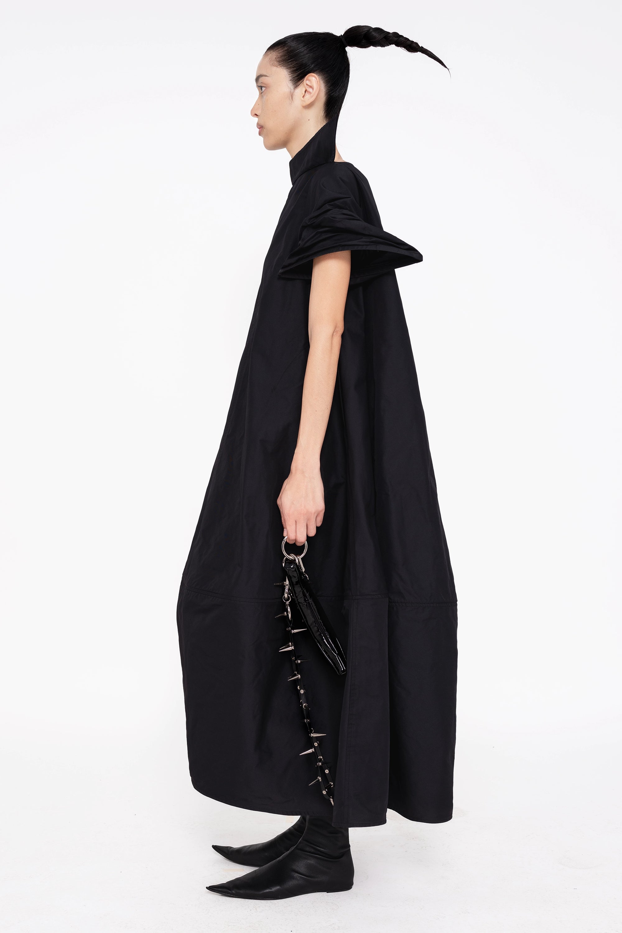 PERSON-WEARING-BLACK-POINTY-SLEEVE-DRESS-HOLDING-BLACK-SPIKED-BANANA-TOTE-WITH-SPIKED-LEATHER-STRAP-AND-BLACK-FLAT-POINTY-TOE-BOOT-SIDE-VIEW