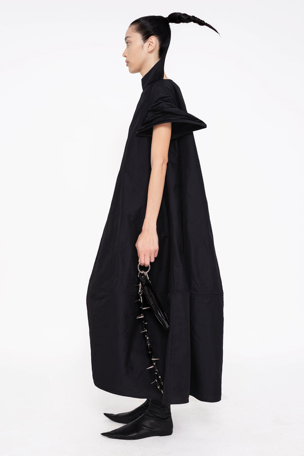 PERSON-WEARING-BLACK-POINTY-SLEEVE-DRESS-HOLDING-BLACK-SPIKED-BANANA-TOTE-WITH-SPIKED-LEATHER-STRAP-AND-BLACK-FLAT-POINTY-TOE-BOOT-SIDE-VIEW