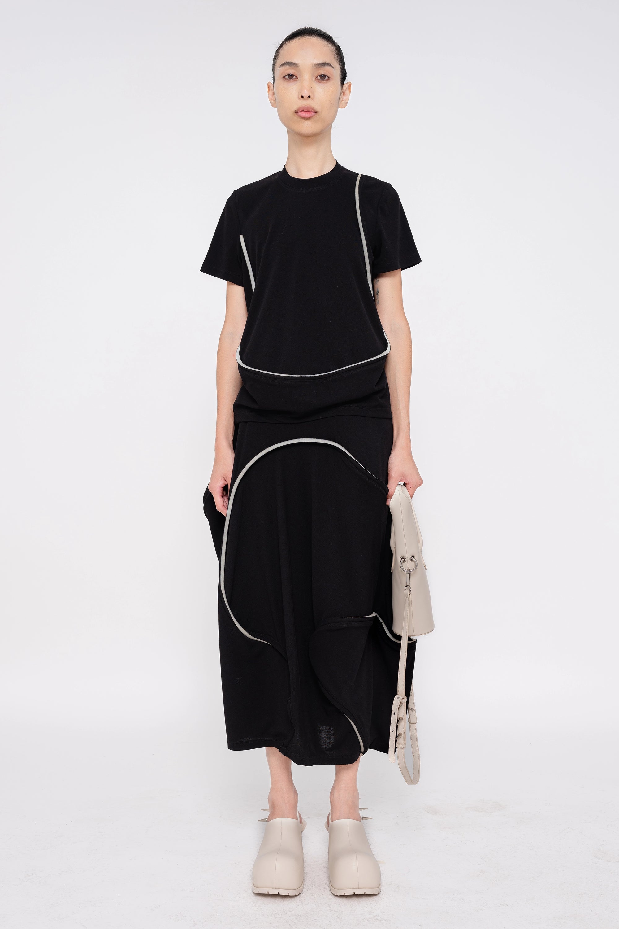 brunette-model-white-background-wearing-black-tee-with-foam-line-detail-along-with-soft-foam-line-detail-while-holding-off-white-pointy-shoulder-bag-and-off-white-spikey-double-heeled-mules-full-body-front