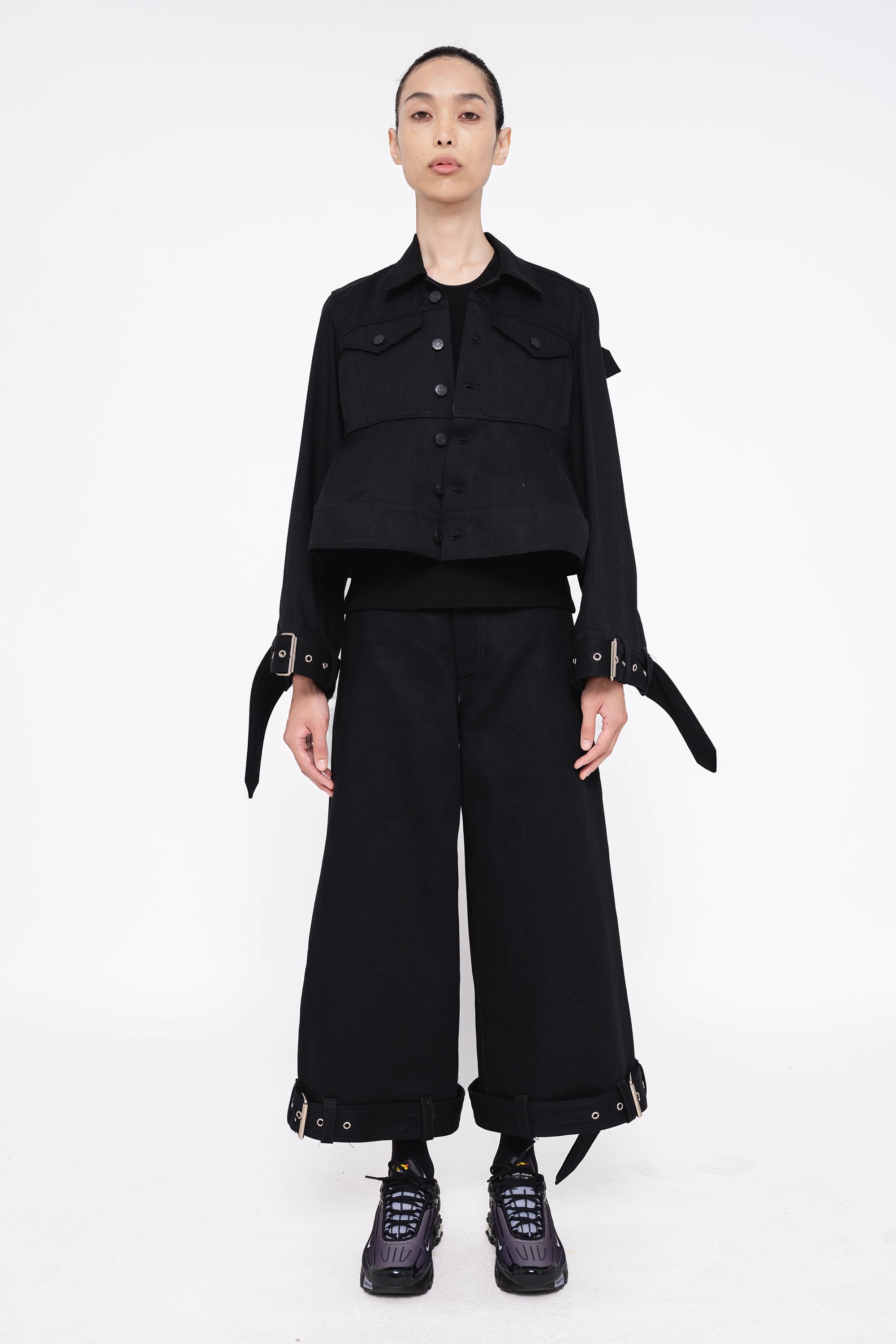 brunette-model-wearing-black-cropped-jean-jacket-with-belted-sleeve-design-also-wearing-black-denim-belted-hem-pants-with-sneakers-full-body-front