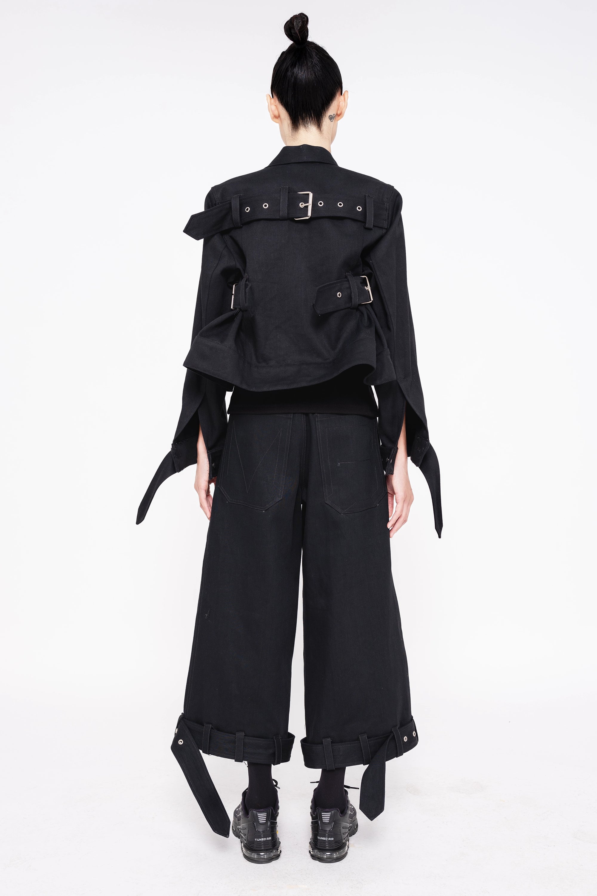 brunette-model-wearing-black-cropped-jean-jacket-with-belted-sleeve-and-back-design-also-wearing-black-denim-belted-hem-pants-with-sneakers-full-body-back