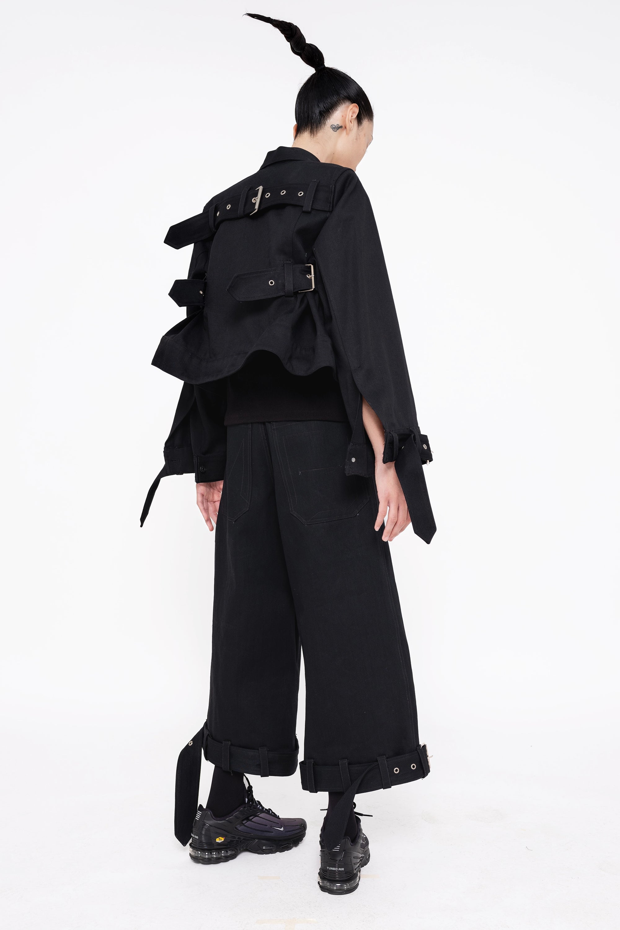 brunette-model-wearing-black-cropped-jean-jacket-with-belted-sleeve-and-back-design-also-wearing-black-denim-belted-hem-pants-with-sneakers-full-body-back