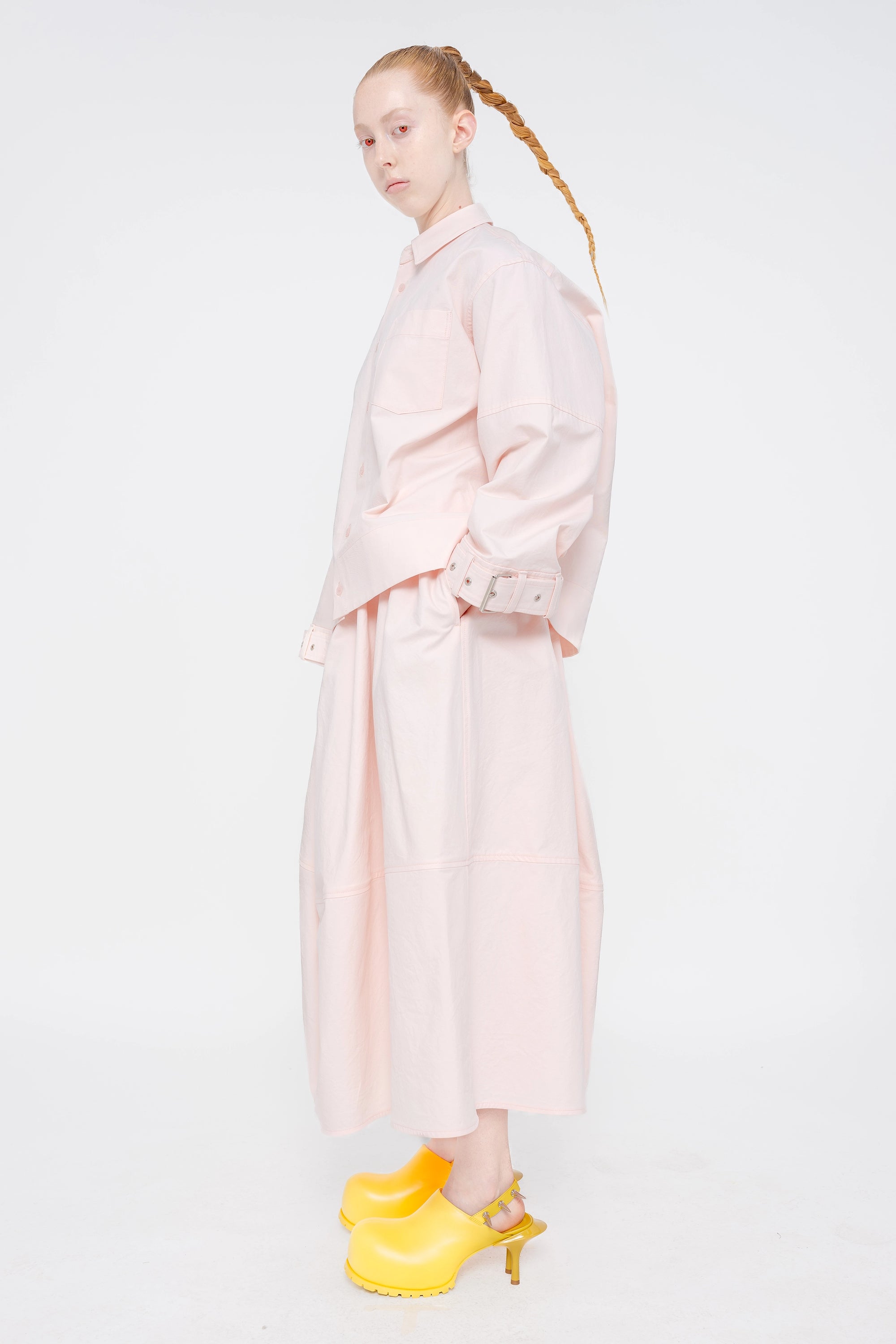 blonde-model-red-eyes-white-background-wearing-soft-pick-shirt-with-belted-sleeve-detail-and-soft-pink-volume-skirt-with-mustard-yellow-spikey-doubled-heeled-mules-full-body-side