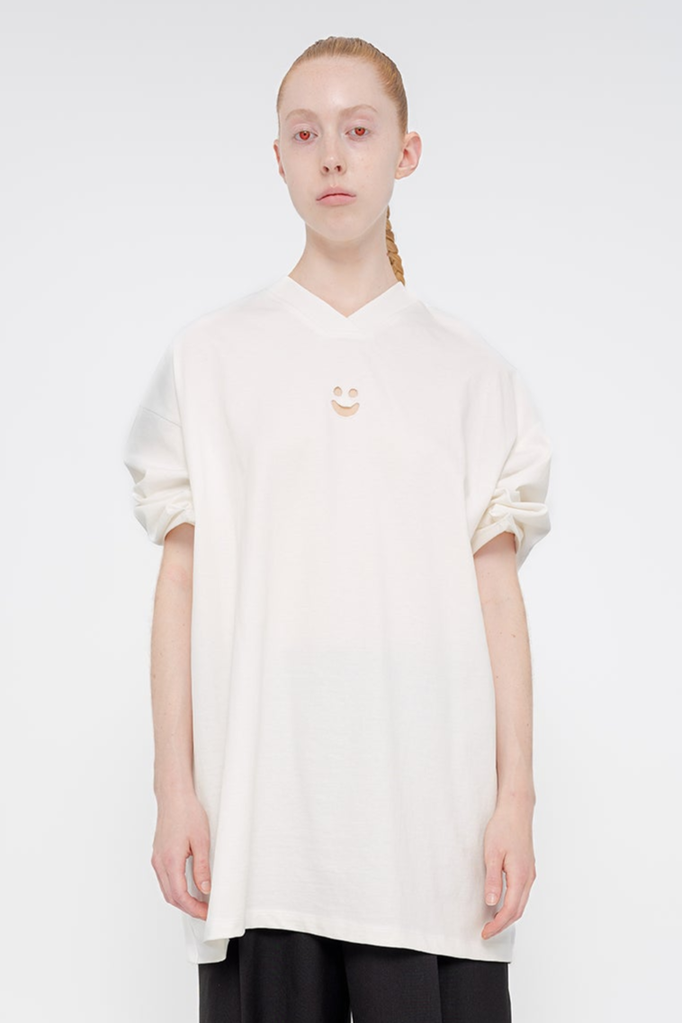 PERSON-WEARING-WHITE-POINTY-SLEEVE-TEE-WITH-SMALL-SMILEY-FACE-CUT-OUT-ON-CHEST-WITH-BLACK-PANTS-CLOSE-UP-FRONT-VIEW