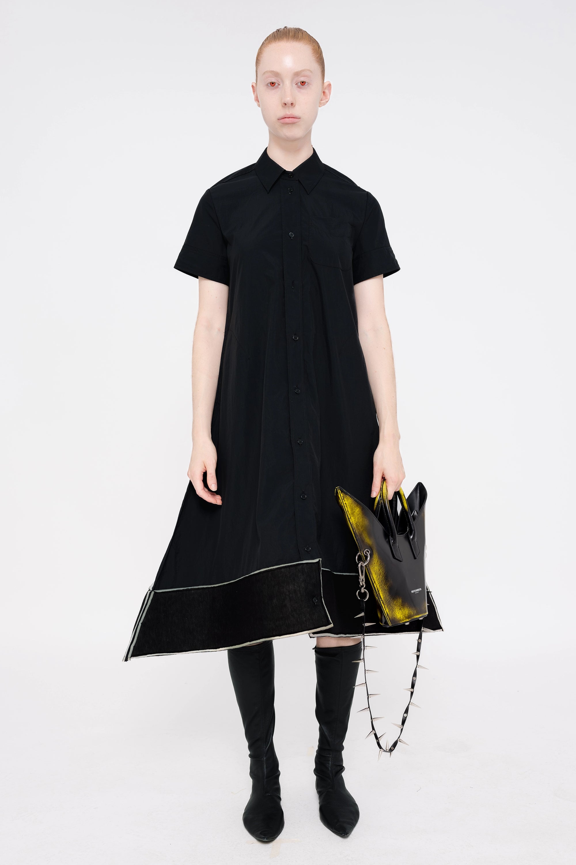 PERSON-WEARING-BLACK-A-LINE-SHORT-SLEEVE-BUTTON-DOWN-DRESS-WITH-FOAM-HEM-DETAIL-WITH-BLACK-FLAT-POINTY-TOE-BOOTS-HOLDING-PAINTED-BLACK-POINTY-SHOULDER-BAG-WITH-SPIKED-LEATHER-STRAP-FRONT-VIEW