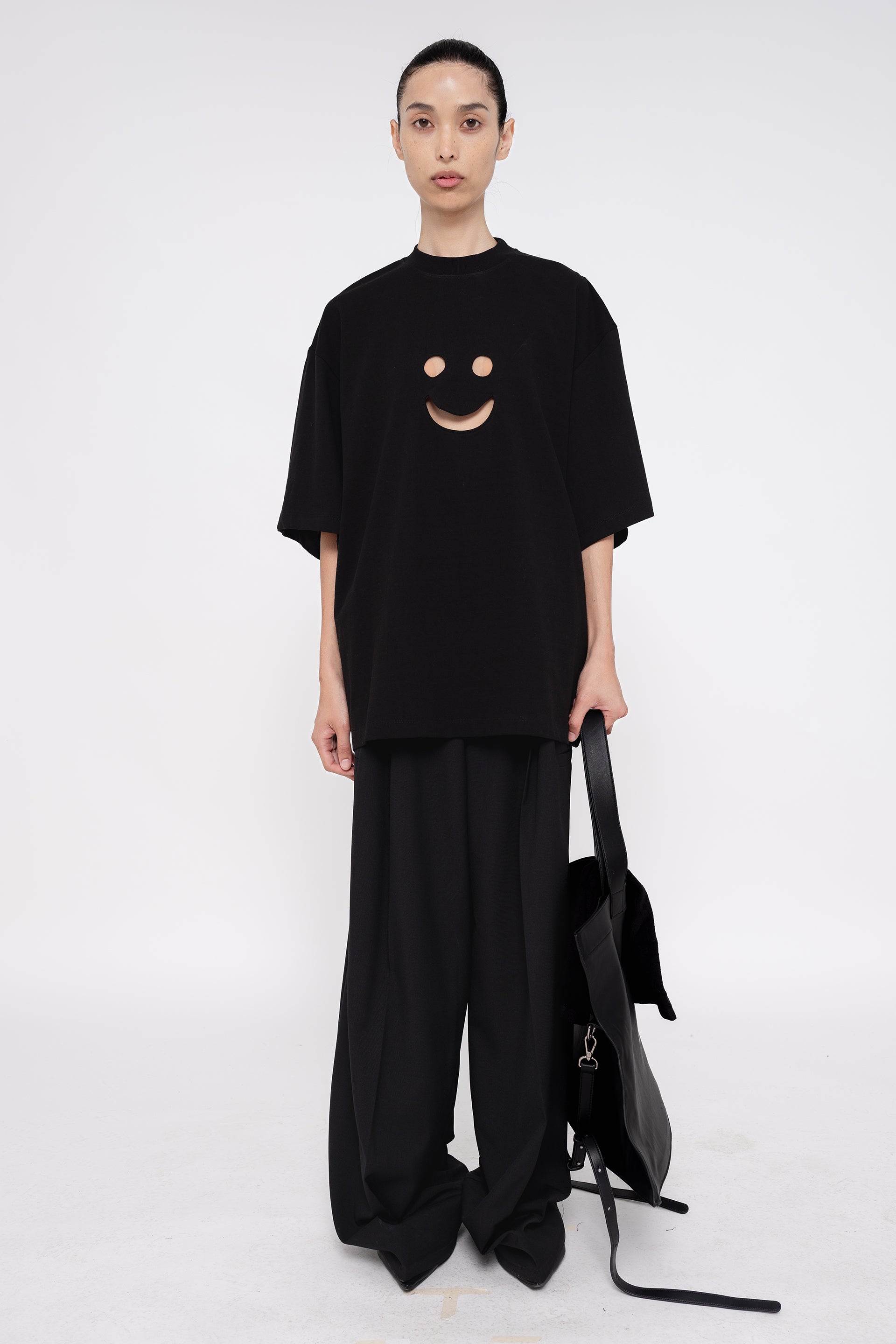 PERSON-WEARING-BLACK-TEE-WITH-SMILEY-FACE-CUT-OUT-AND-BLACK-TAILORED-PANTS-HOLDING-BLACK-SLOUCHY-WEEKENDER-BAG-WITH-POINTY-BLACK-BOOTS-FRONT-VIEW