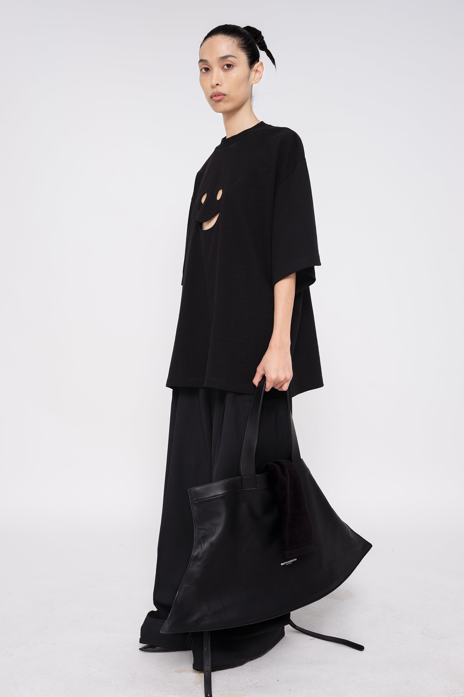 PERSON-WEARING-BLACK-TEE-WITH-SMILEY-FACE-CUT-OUT-AND-BLACK-TAILORED-PANTS-HOLDING-BLACK-SLOUCHY-WEEKENDER-BAG-WITH-POINTY-BLACK-BOOTS-THREE-QUARTER-VIEW