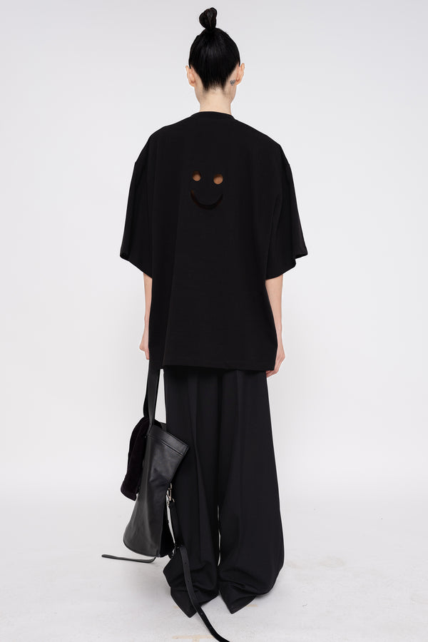 PERSON-WEARING-BLACK-TEE-WITH-SMILEY-FACE-CUT-OUT-AND-BLACK-TAILORED-PANTS-HOLDING-BLACK-SLOUCHY-WEEKENDER-BAG-WITH-POINTY-BLACK-BOOTS-BACK-VIEW