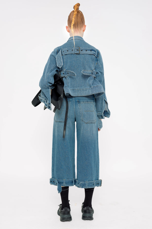 blonde-model-white-background-wearing-medium-wash-jeans-and-jean-jacket-with-belted-hem-detail-model-is-holding-black-bag-and-wearing-black-socks-and-nike-sneakers-full-body-back