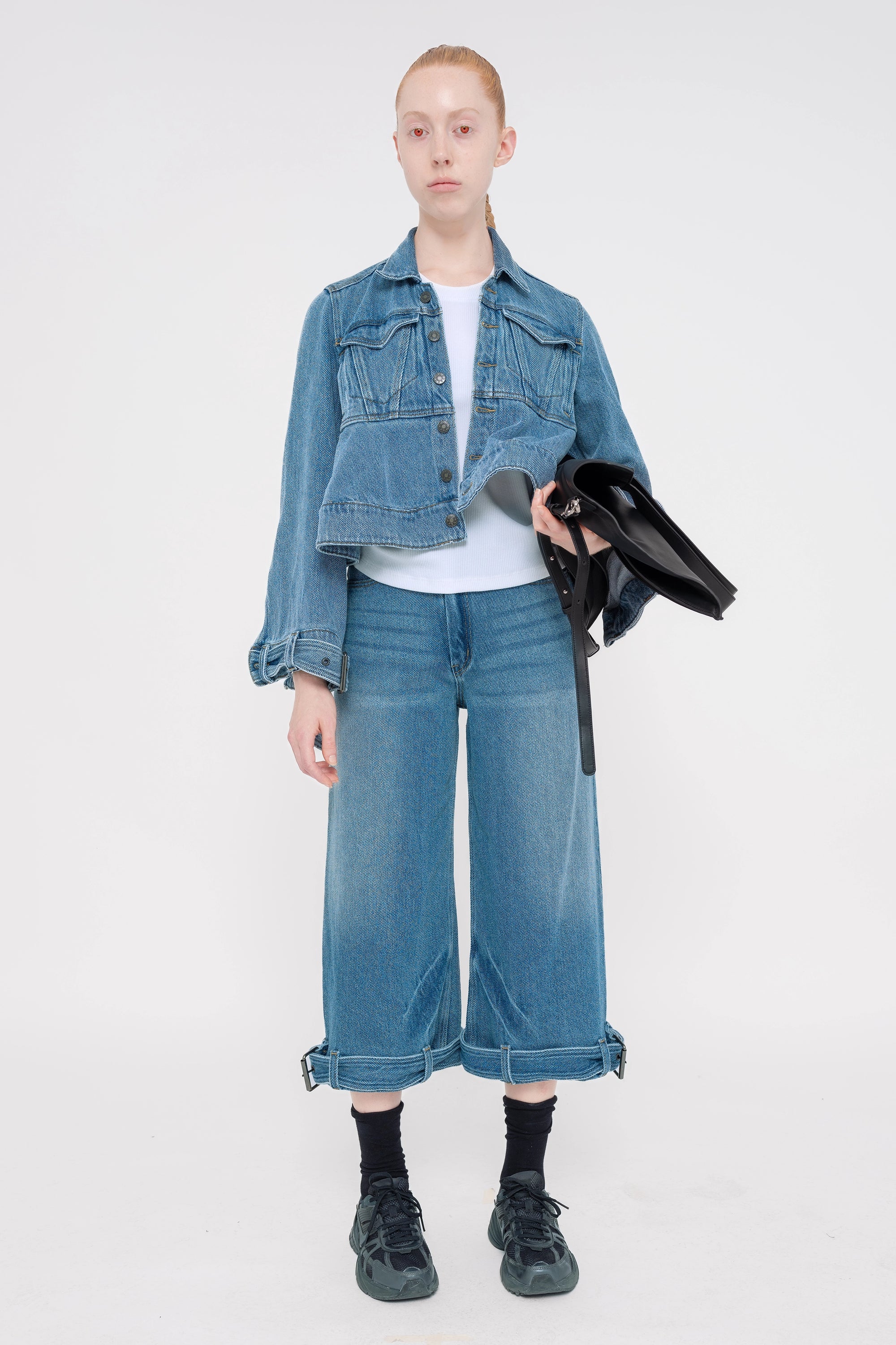 blonde-model-white-background-wearing-medium-wash-jeans-and-jean-jacket-with-belted-hem-detail-model-has-red-eyes-holding-black-bag-and-wearing-black-socks-and-nike-sneakers-full-body-front