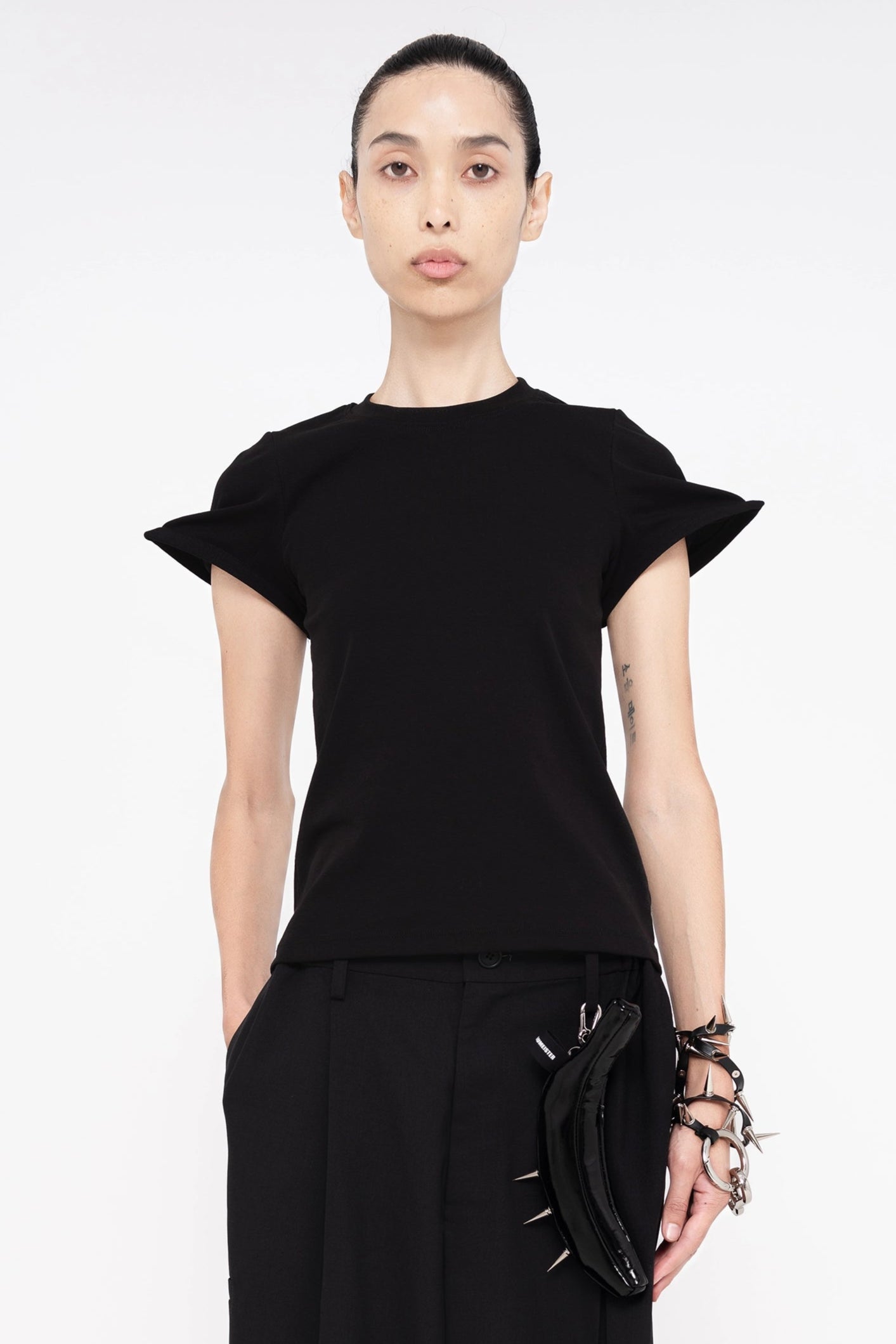 PERSON-WEARING-BLACK-POINTY-SLEEVE-TEEWITH-BLACK-TAILORED-PANTS-ACCESSORIZED-WITH-BLACK-SPIKED-BANANA-TOTE-AND-SPIKED-LEATHER-CHAIN-ON-WRIST-CLOSE-UP-FRONT-VIEW