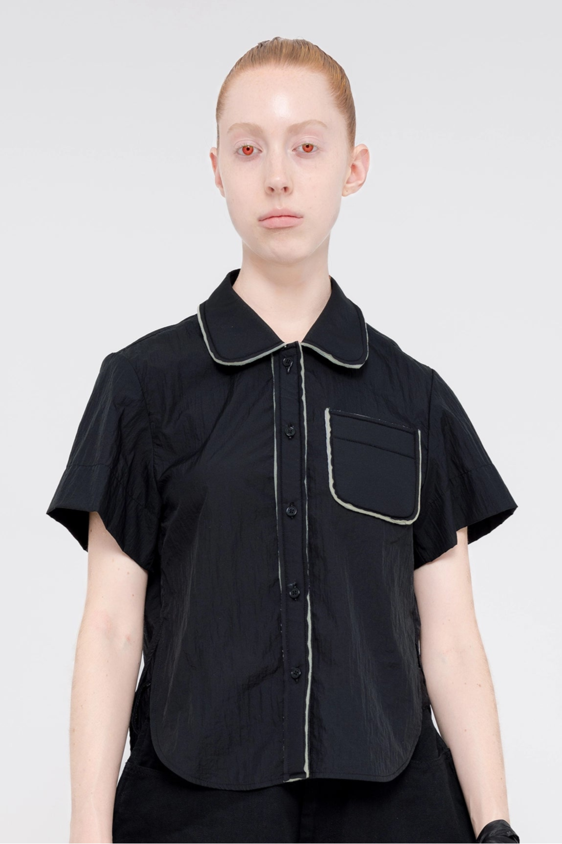 blonde-model-white-background-red-eyes-wearing-cropped-short-sleeve-black-button-down-with-foam-detail-on-collar-button-area-and-pocket-quarter-view-front-facing