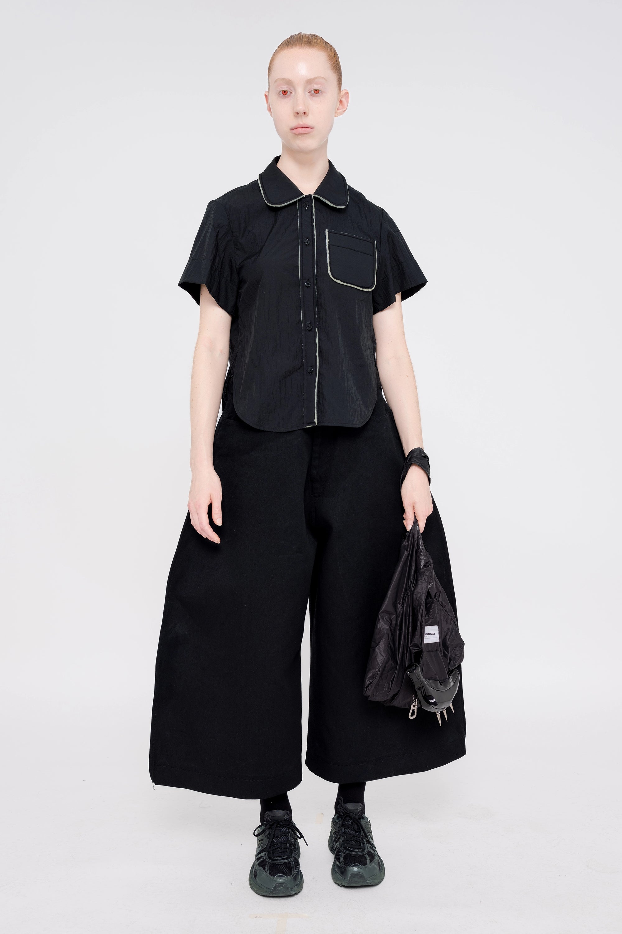 blonde-model-white-background-red-eyes-wearing-cropped-short-sleeve-black-button-down-with-foam-detail-on-collar-button-area-and-pocket-wide-leg-pants-with-glossy-banana-tote-and-sneakers-full-body-front