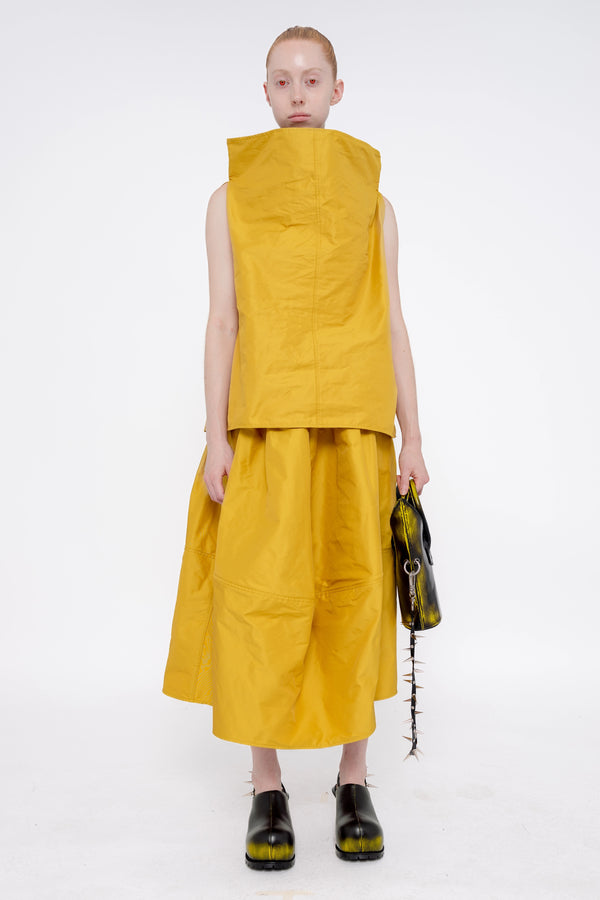 blonde-female-model-white-background-mustard-yellow-boatneck-top-and-skirt-red-eyes-holding-painted-pointed bag-with-spikey-strap-and-black-painted-spikey-doubled-heeled-mules-front-full-body