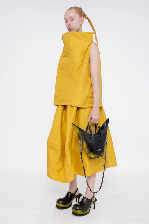 blonde-female-model-white-background-mustard-yellow-boatneck-top-and-skirt-red-eyes-holding-painted-pointed bag-with-spikey-strap-and-black-painted-spikey-doubled-heeled-mules-full-body-side