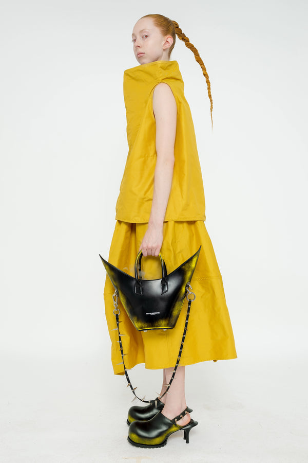 blonde-female-model-white-background-mustard-yellow-boatneck-top-and-skirt-red-eyes-holding-painted-pointed bag-with-spikey-strap-and-black-painted-spikey-doubled-heeled-mules-full-body-side