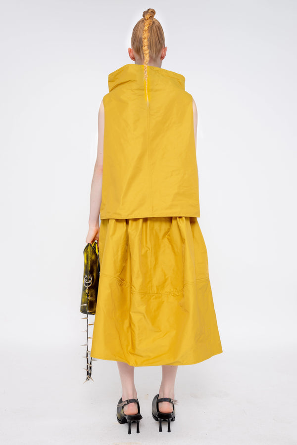 blonde-female-model-white-background-mustard-yellow-boatneck-top-and-skirt-holding-painted-pointed bag-with-spikey-strap-and-black-painted-spikey-doubled-heeled-mules-full-body-back