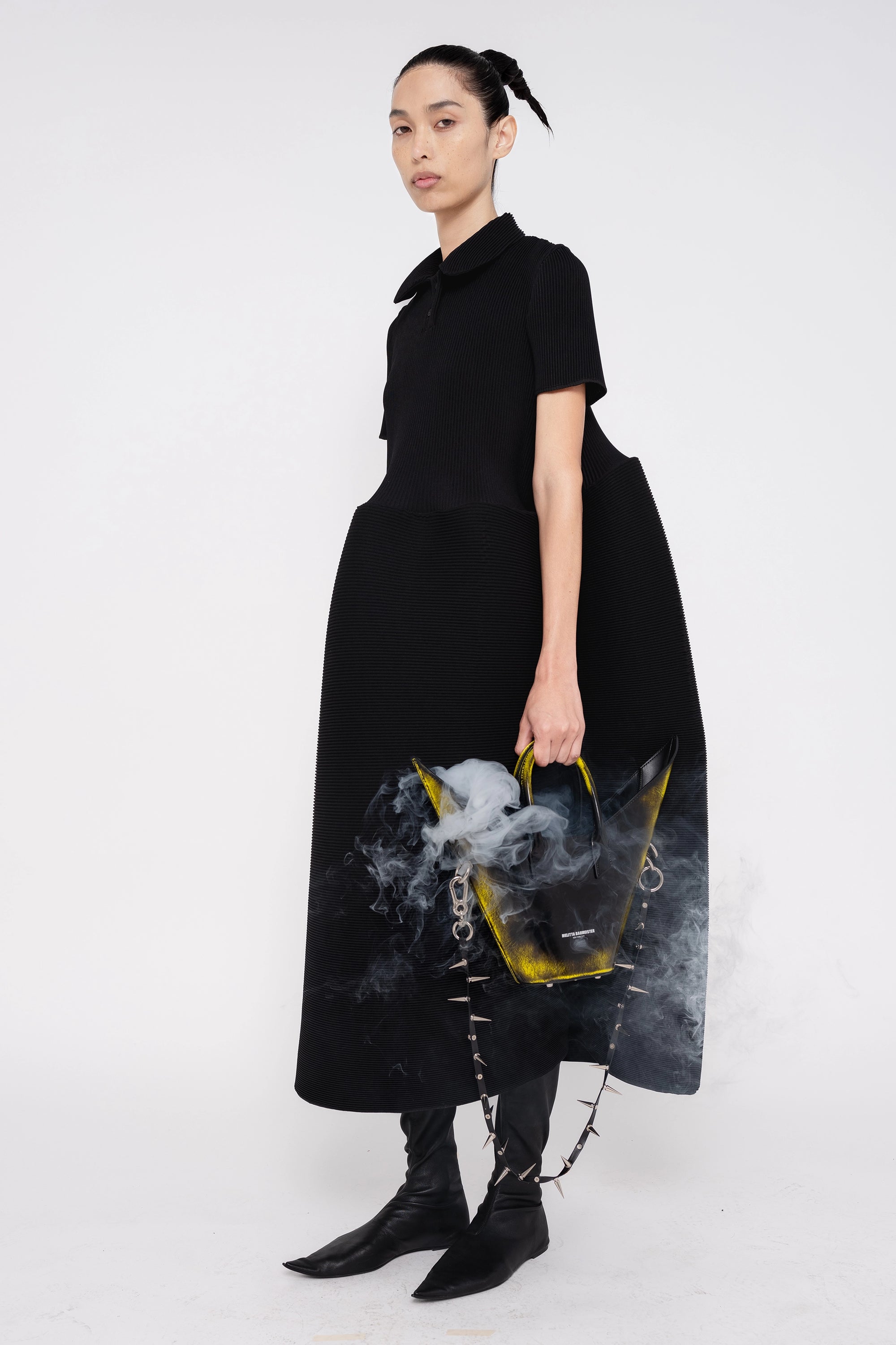 PERSON-WEARING-BLACK-RIPPLE-PLEAT-POLO-DRESS-WITH-BLACK-FLAT-POINTY-BOOTS-HOLDING-BLACK-PAINTED-POINTY-SHOULDER-BAG-WITH-SPIKED-CROSSBODY-STRAP-WITH-SMOKE-COMING-OUT-OF-BAG-THREE-QUARTER-VIEW