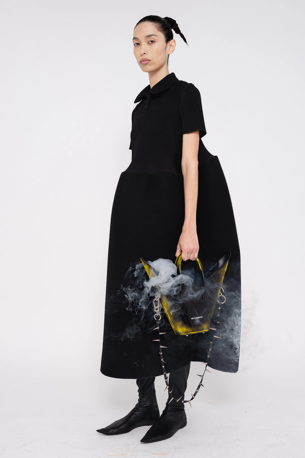 PERSON-WEARING-BLACK-RIPPLE-PLEAT-POLO-DRESS-WITH-BLACK-FLAT-POINTY-BOOTS-HOLDING-BLACK-PAINTED-POINTY-SHOULDER-BAG-WITH-SPIKED-CROSSBODY-STRAP-WITH-SMOKE-COMING-OUT-OF-BAG-THREE-QUARTER-VIEW