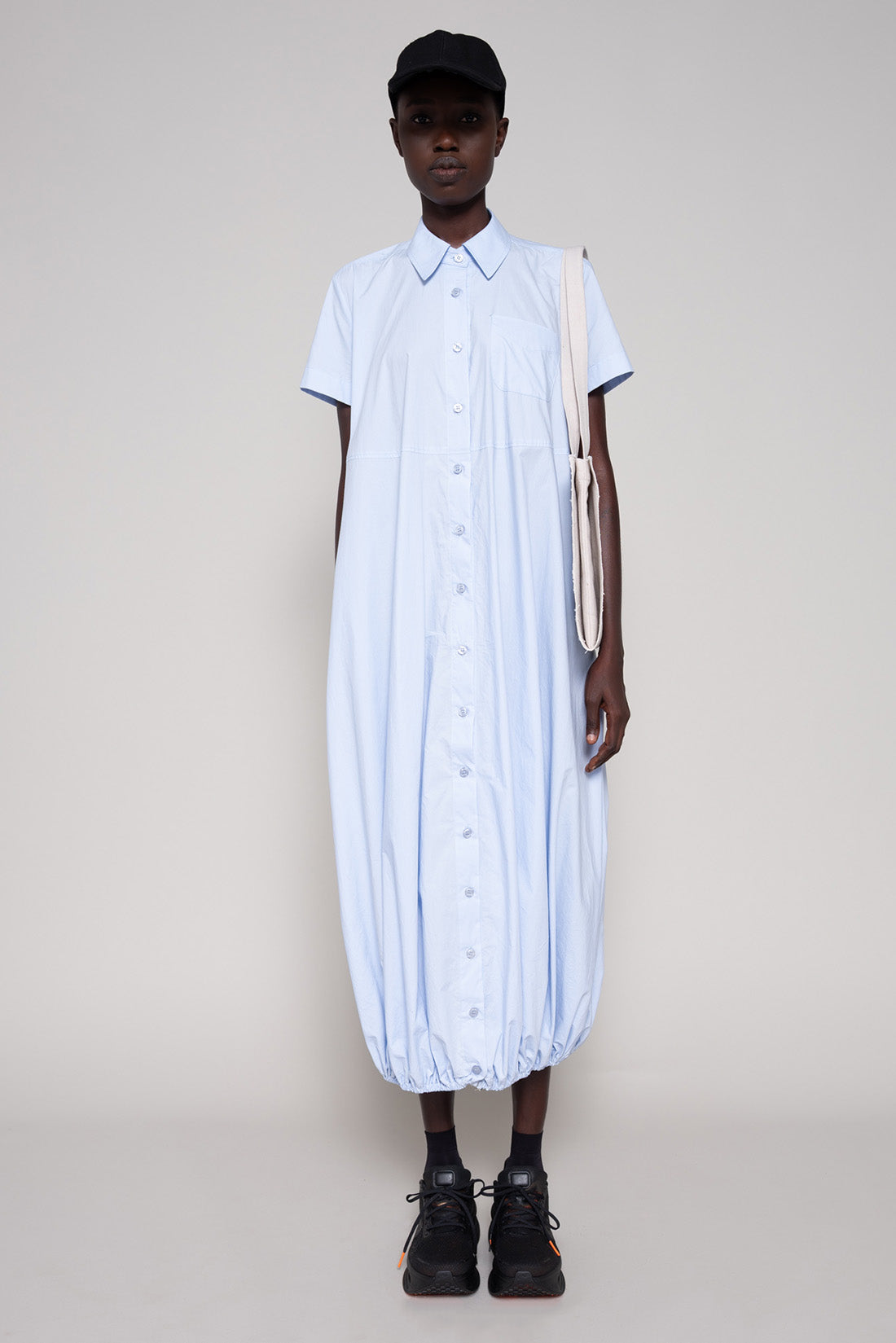 Person wearing a light blue dress with a black cap against a plain background