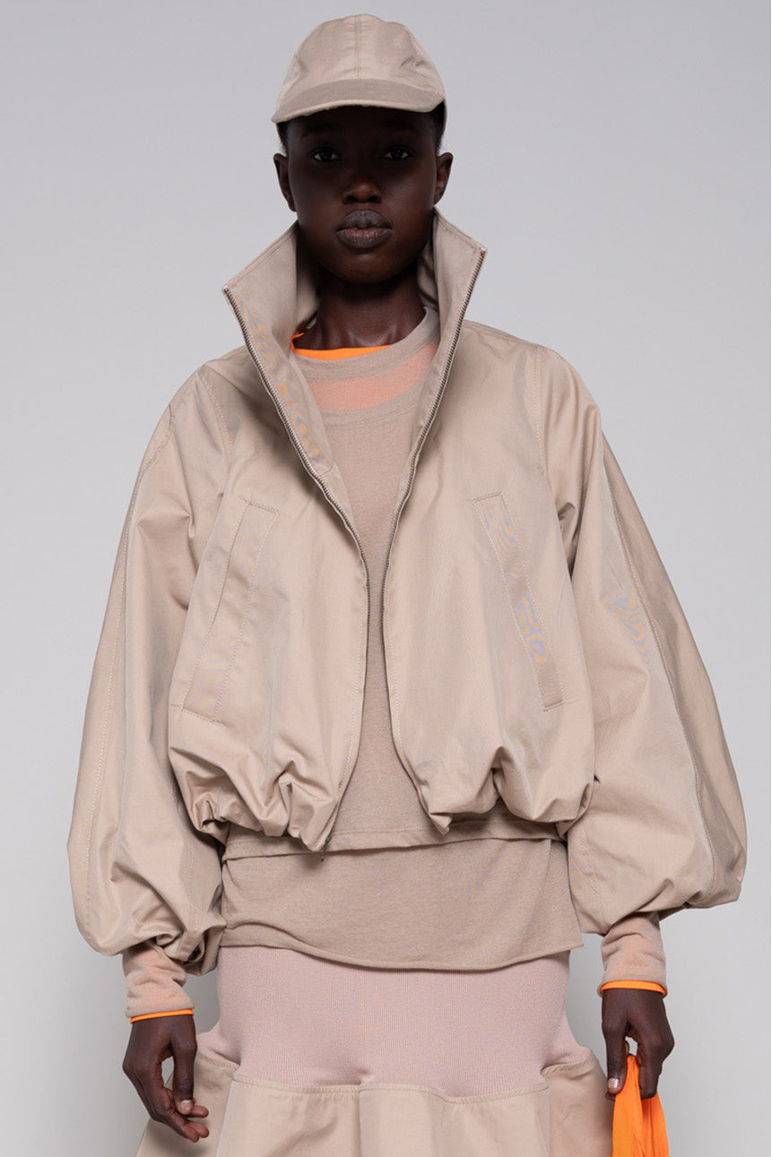 Person wearing a beige top,  jacket, skirt and cap with an orange tote bag and black sneakers against a plain background