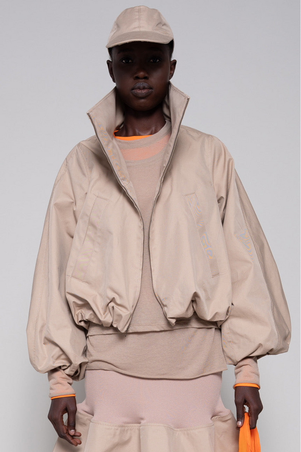 Person wearing a beige top,  jacket, skirt and cap with an orange tote bag and black sneakers against a plain background