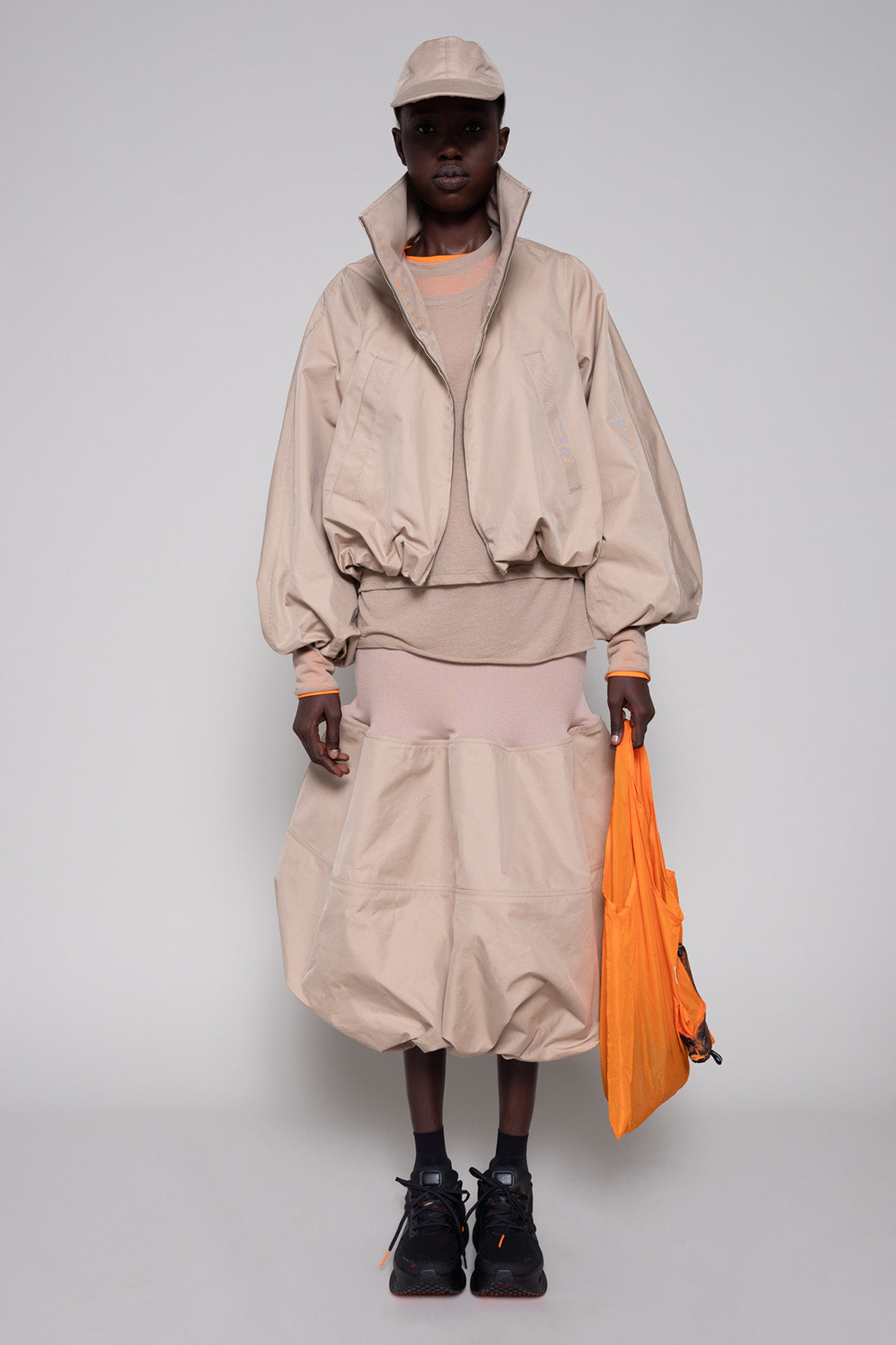 Person wearing a beige top, jacket, skirt and cap with an orange tote bag and black sneakers against a plain background