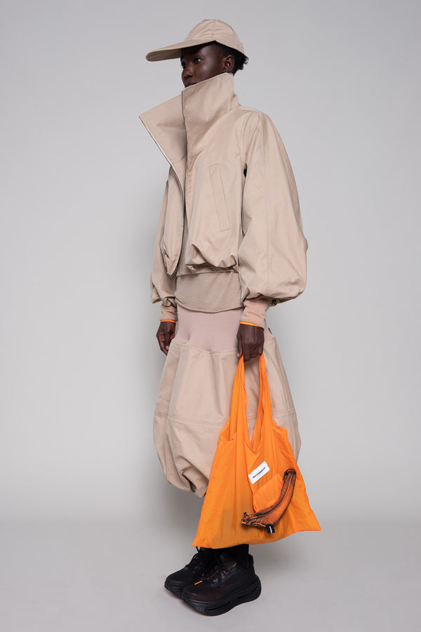 Person wearing a beige jacket, skirt and cap with an orange tote bag and black sneakers against a plain background