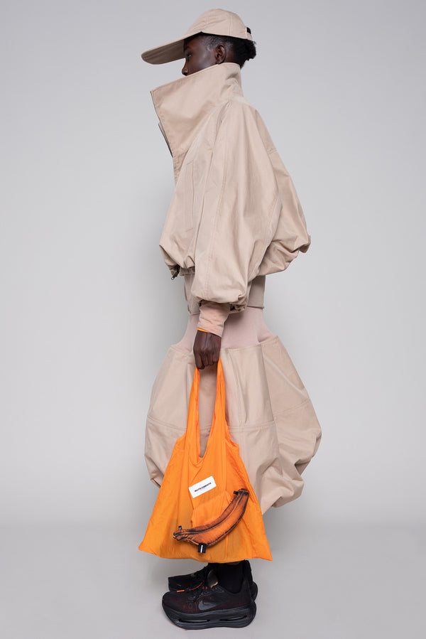 Person wearing a beige jacket, skirt and cap with an orange tote bag and black sneakers against a plain background